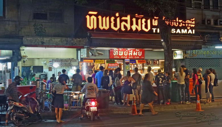 Hàng người xếp chờ trước cửa Thip Samai buổi tối, ánh đèn vàng, biển hiệu đỏ đặc trưng